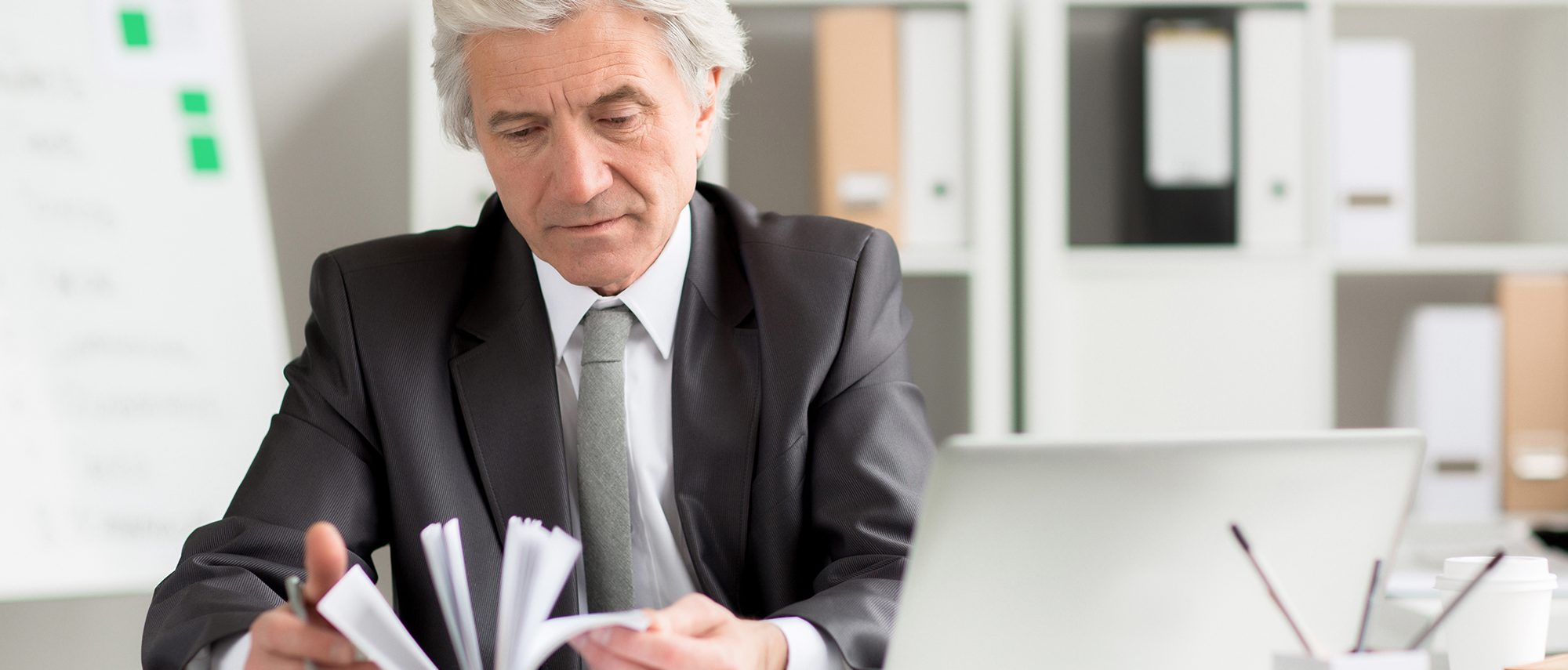 Grey-haired businessman in formalwear looking for some specific notes in his notebook by workplace
